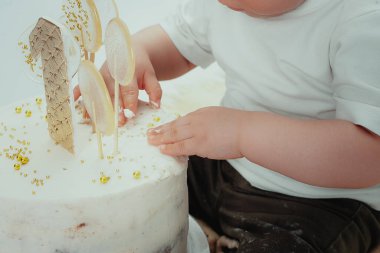 Küçük çocuk beyaz pastanın yanında oturuyor, elleriyle dokunuyor ve tatmaya çalışıyor. Çocuğun pastanın kremasından kirli elleri ve pantolonu var. Hayatın tatlı bir yılı. Tasasız çocukluk.
