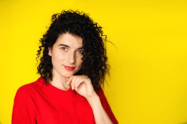Sarı arka planda kırmızı tişörtlü kıvırcık siyah saçlı çekici bir genç kız. Portre fotoğrafçılığı. Duygular, yüz ifadeleri. Kıvırcık, esmer. Kırmızı ruj.