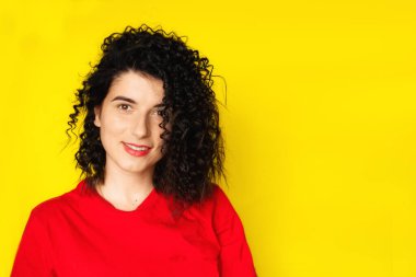 Sarı arka planda kırmızı tişörtlü kıvırcık siyah saçlı çekici bir genç kız. Portre fotoğrafçılığı. Duygular, yüz ifadeleri. Kıvırcık, esmer. Kırmızı ruj.
