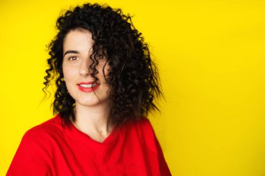 Sarı arka planda kırmızı tişörtlü kıvırcık siyah saçlı çekici bir genç kız. Portre fotoğrafçılığı. Duygular, yüz ifadeleri. Kıvırcık, esmer. Kırmızı ruj.