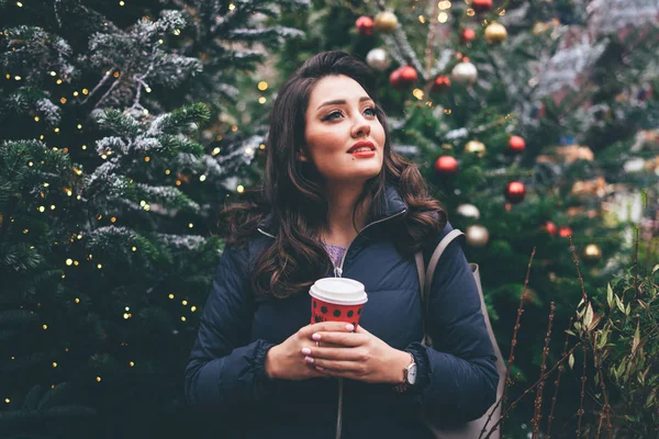Young woman with makeup with paper cup near Christmas trees looking up.