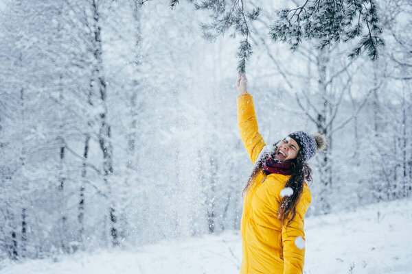 Young girl in down jacket and hat in winter forest shakes a snowy branch.