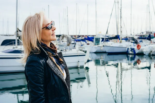 Portrait of an adult beautiful lady in a jacket standing in a marina and looks into the distance in sunglasses  on yachts background.