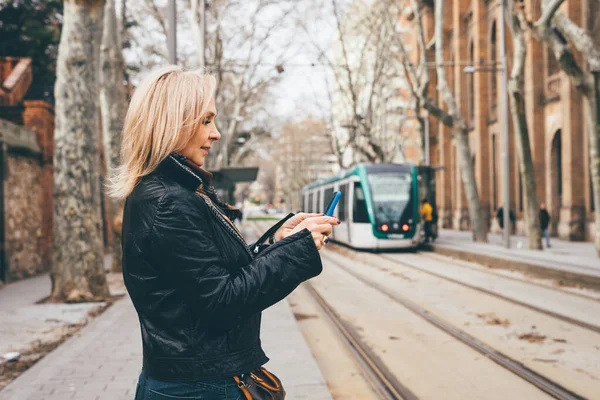 An adult beautiful lady in a jacket stands near the tram of Barcelona and looks at the phone. View assembly close.