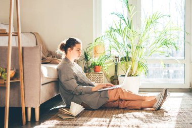 Evden çalışan genç bir kadın. Kız güneşli odada dizüstü bilgisayar kullanıyor..