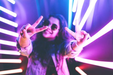 Neşeli, sevimli, gülümseyen kız neon arka planda barış işareti gösteriyor..