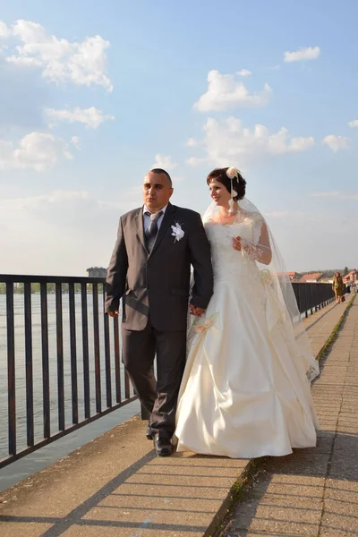 The bride and groom in a white dress walk along the embankment of the river in sunny weather and talk about the future