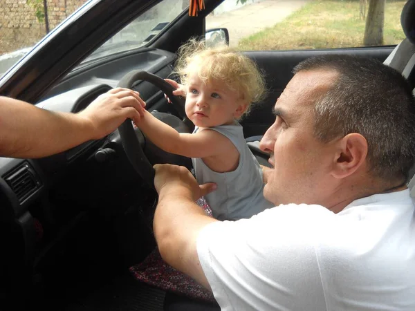 A small child is driving a car. A middle-aged father supports him with his hands. The boy plays in the driver of the car.