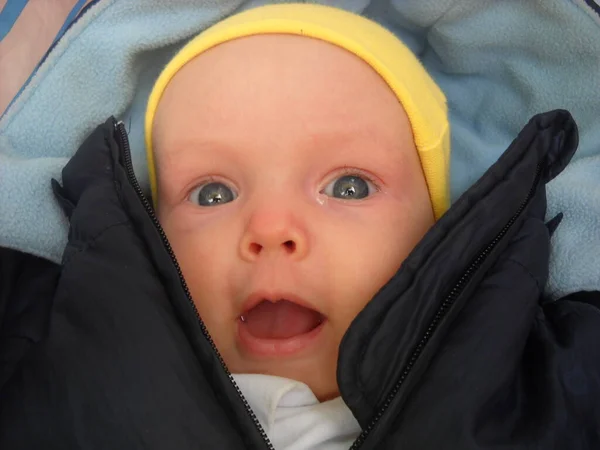 아기는 유모차에 누워서 깜짝 놀라 입을 벌린다. 어린이는 가벼운 모자를 쓰고 따뜻 한 점프수트를 입고 있다. 왼쪽 눈에서 눈물이 흘러나온다 — 스톡 사진