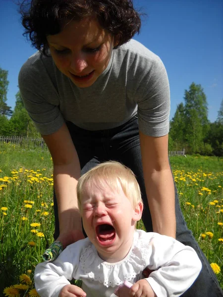 Ein kleines Kind weint sehr. Mutter versucht ihn mit den Händen zu halten und den Wutanfall zu stoppen. Das Kind ist nicht glücklich. Er trägt ein weißes Hemd. Im Hintergrund eine ländliche Landschaft mit grünem Gras — Stockfoto