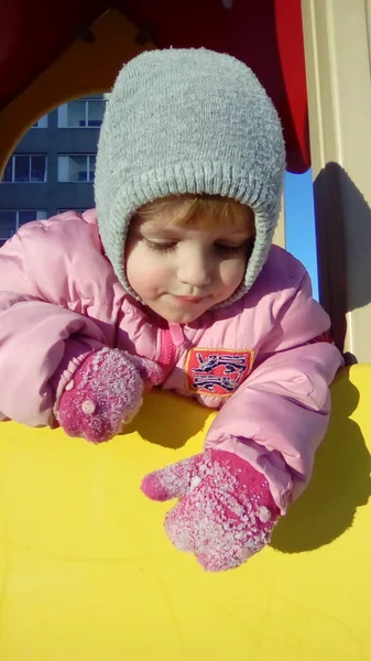 Una niña cuelga de un tobogán de madera y mira hacia abajo. El niño lleva un sombrero gris, una chaqueta rosa gruesa, mitones de frambuesa con hielo y bolas de nieve. Clima helado soleado —  Fotos de Stock