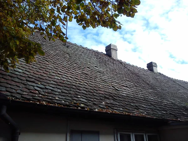 El techo de la casa con azulejos viejos. Un edificio de piedra con baldosas de arcilla quemada. Día soleado en otoño. Plátano alto con hojas que caen amarillentas. Las tejas están cubiertas de musgo y hojas —  Fotos de Stock