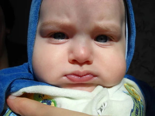 Le petit enfant boudait et fronçait les sourcils. Fatigué ou endormi. Sur la tête des garçons se trouve une capuche d'une combinaison en peluche bleue. Lumière naturelle du soleil par la fenêtre . — Photo