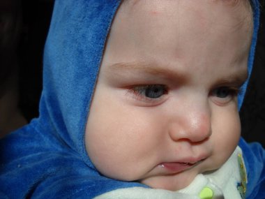 Küçük bir çocuk kaşlarını çatar ve kaşlarını çatar. Bir yana bak. Çocuğun kafasında mavi pelüş bir kapüşon var. Kaputun altına saklanmış saçlar, temiz geniş bir alın. Pencereden gelen doğal güneş ışığı.