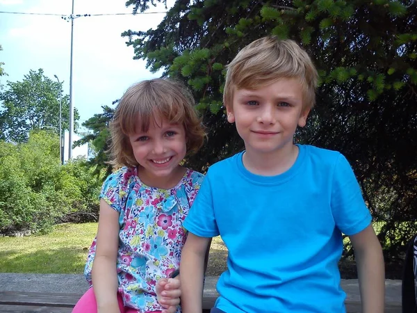 Un niño y una niña con el pelo rubio, la piel clara y los ojos azules están sentados uno al lado del otro y sonriendo, mirando a la cámara. Los niños usan ropa de verano. En el fondo hay árboles y arbustos . — Foto de Stock