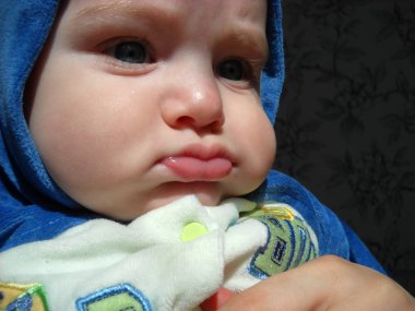 Baby 6 months old on mother's hands. The boy is dressed in a jumpsuit and a blue hood. A child with thick cheeks and a drooping lower lip. Looking to the side, head turning three quarters clipart