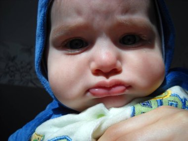 Küçük çocuk somurttu ve kaşlarını çattı. Kafasına mavi peluş bir tulum geçirildi. Çocuğun kalın yanakları ve gücenmiş bir ifadesi var. Bebek daha 6 aylık. Annem bir çocuğu kollarında tutuyor.