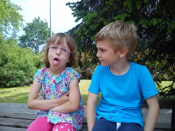 La fille avec des lunettes montre la langue, le garçon dans un T-shirt bleu regarde la fille. Frère et sœur sont assis sur un banc au milieu d'une pelouse verte, des buissons et des arbres. Vacances scolaires d'été dans la ville — Photo