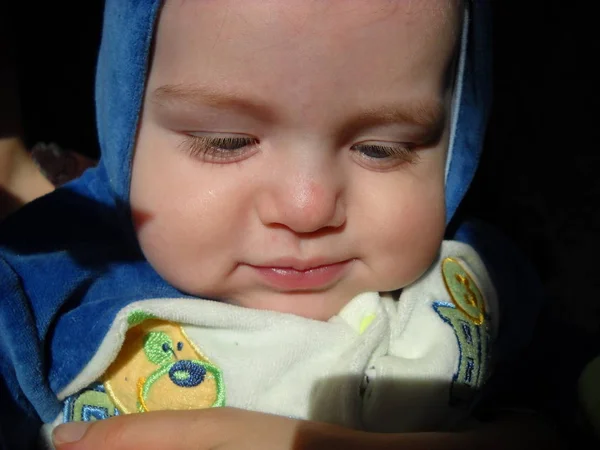 Χαριτωμένο μωράκι 6 μηνών με κουκούλα από μπλε φόρμα. Το αγόρι κοιτάζει κάτω και χαμογελάει ντροπαλά. Η φυσική όψη του δέρματος, οι φωτεινές βλεφαρίδες είναι αξιοσημείωτες. Υψηλό μέτωπο — Φωτογραφία Αρχείου