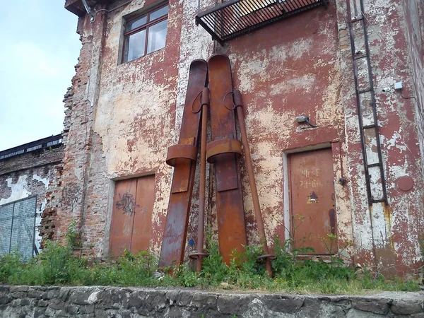 Huge skis on the facade of the old factory. Skis and ski poles are made of rusty metal and lean against the wall. Abandoned factory. Oxidation of iron. The shabby facade of the building — Stock Photo, Image