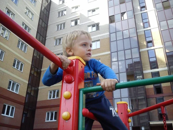 Moscú, Rusia - 20 de agosto de 2019 - un chico lindo con el pelo rubio trepó en un torniquete de niños. Parque infantil en la zona de los nuevos edificios. Edificio de ladrillo de gran altura con fachada de vidrio —  Fotos de Stock