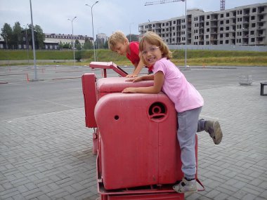 Çocuklar sokağa açıkta kalan eski fabrika makinesine tırmanıyorlar. Fabrika traktör-makine pembe boya. Gülen kız ciddidir. Sarı saçlı çocuklar. Arkaplanda inşaat