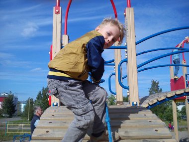 Çocuk parkında. Sarı saçlı bir çocuk. Rüzgarda dalgalanan saçlar. Çocuk bir çocuk kaydırağından atlamaya hazır. İnsanlar parkta yürüyor.