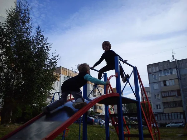 Junge und Mädchen, Bruder und Schwester spielen auf dem Spielplatz. Kinder kletterten auf eine Metallrutsche. das Mädchen erhob sich. klettert der Junge die Metalloberfläche hinauf. Im Hintergrund ein Innenhof der Stadt — Stockfoto