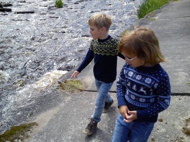 Erkek ve kız kardeş nehrin yanında yürüyorlar. Oğlan ve kız mavi kot pantolon ve mavi örülmüş kalın kazaklar ve yeni yıl süslemeleri giyiyorlar. Yazın başında. Konuşma ve oyun