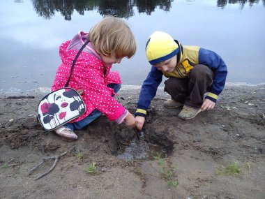 Erkek ve kız kardeş nehrin yanında yürüyorlar. Bir erkek ve bir kız kot pantolon ve sonbahar kıyafetleri giyerler. Yazın başında. Konuşun ve su kenarında oynayın. Beyaz benekli pembe pelerin, sarı ceket.
