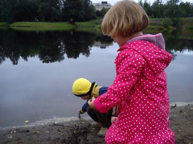 Erkek ve kız kardeş nehrin yanında yürüyorlar. Bir erkek ve bir kız kot pantolon ve sonbahar kıyafetleri giyerler. Yazın başında. Konuşun ve su kenarında oynayın. Beyaz benekli pembe pelerin, sarı ceket.