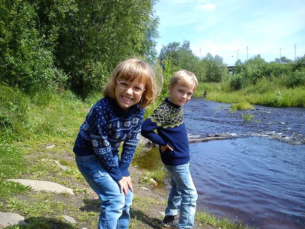 Hermano y hermana están caminando cerca del río. El niño y la niña están vestidos con vaqueros azules y suéteres y jerséis calientes de punto azul con adornos de Año Nuevo. A principios del verano. Conversación y juego —  Fotos de Stock