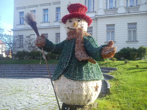 Snowman in a city park. Red hat, green jacket and a broom in the hand of a snowman. Sculpture for evening street lighting. Garland. — Stock Photo, Image