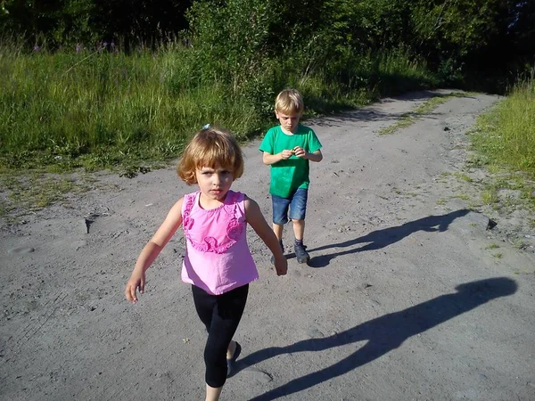 Blondhaarige Kinder laufen im Sommer auf einem Feldweg. Junge in grünem T-Shirt. Mädchen in einer rosafarbenen Bluse mit Rüschen. Bruder und Schwester eilen nach Hause. Wandern in der Natur bei sonnigem Wetter. Schwungvolle Bewegungen. — Stockfoto
