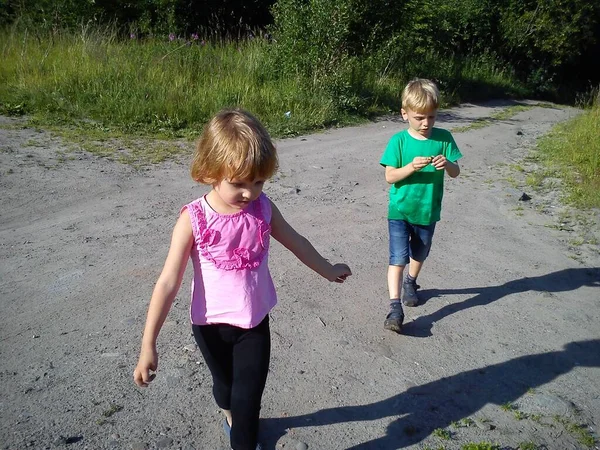 금발의 아이들은 여름에 더러운 길을 걷습니다. 녹색 티셔츠를 입은 소년. 핑크 블라우스를 입고 프릴을 가진 소녀. 형 과 누나는 집으로 가요. 맑은 날씨에 자연 속을 하이킹하는 것. 전면적 인 움직임. — 스톡 사진