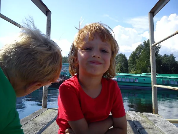 Jungen und Mädchen posieren auf einem Steg. rote und grüne T-Shirts ohne Muster. Kinder spielen in der Nähe von Metallkonstruktionen im Hafen. Schiffe und Wasserfahrzeuge im Hintergrund. Nordsommer. Bruder und Schwester. — Stockfoto