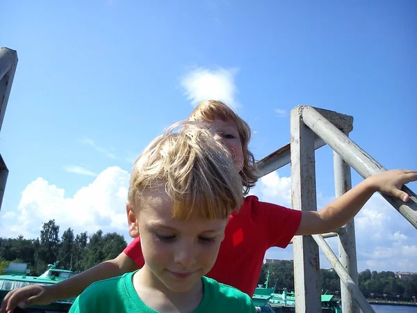Jungen und Mädchen posieren auf einem Steg. rote und grüne T-Shirts ohne Muster. Kinder spielen in der Nähe von Metallkonstruktionen im Hafen. Schiffe und Wasserfahrzeuge im Hintergrund. Nordsommer. Bruder und Schwester. — Stockfoto