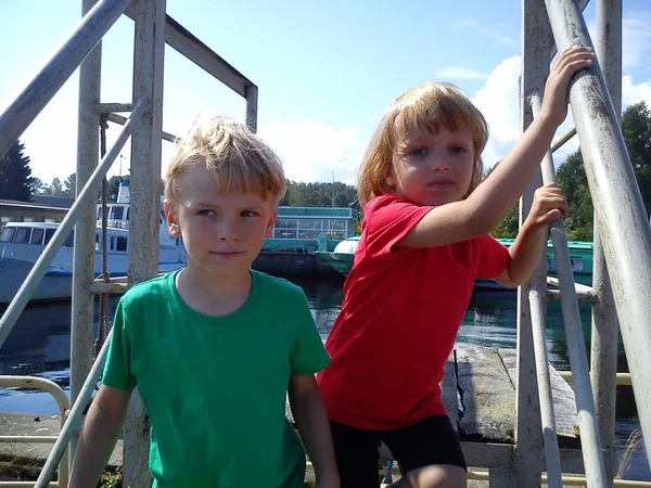 Jungen und Mädchen posieren auf einem Steg. rote und grüne T-Shirts ohne Muster. Kinder spielen in der Nähe von Metallkonstruktionen im Hafen. Schiffe und Wasserfahrzeuge im Hintergrund. Nordsommer. Bruder und Schwester. — Stockfoto