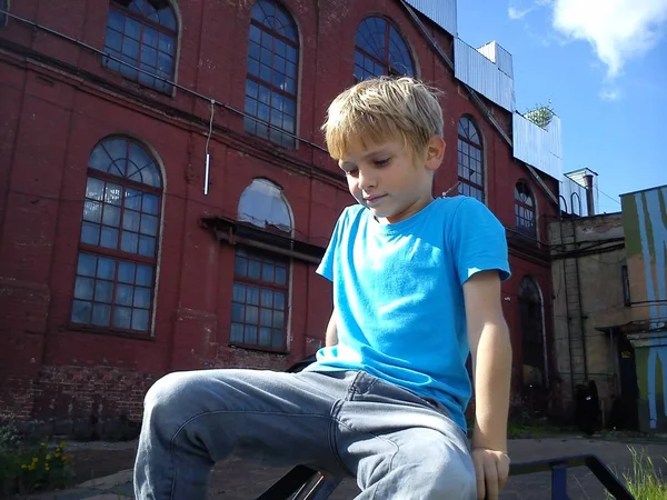 Blonďatý chlapec v modrém tričku a šedých kalhotách si hraje na ulici vedle staré továrny. Dítě na něco vylezlo a podívalo se dolů. V pozadí stará cihlová budova a nebe — Stock fotografie