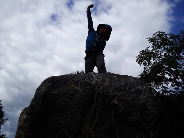 El adolescente trepó sobre una piedra alta y posa. Silueta negra de un deportista sobre un fondo de cielo azul y nubes blancas. Postura desafiante. Llevaba una capucha en la cabeza. Demostración de agilidad — Foto de Stock