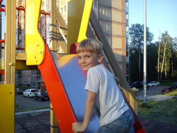 Un niño con el pelo rubio subió una colina en el patio de recreo, se vuelve y sonríe. Equipo de juego para la calle. Pinturas rojas y amarillas brillantes en las paredes de la zona infantil . —  Fotos de Stock