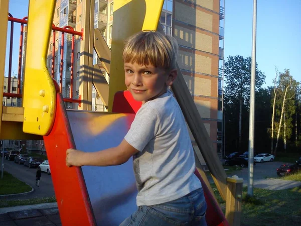 Um menino com cabelo loiro subiu uma colina no playground, volta atrás, sorri e franze a testa. Equipamento de jogos para a rua. Cores vermelhas e amarelas brilhantes . — Fotografia de Stock