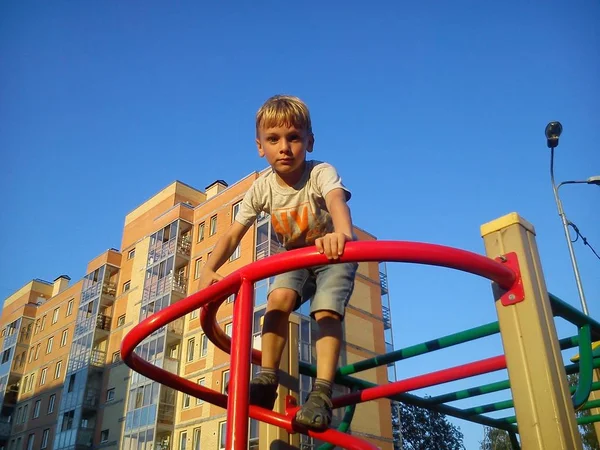 Blonďatý chlapec se vyšplhal po kovové konstrukci na hřišti, rukama se chytil zábradlí a podíval se dolů. Herní zařízení pro dvorek. Cihlový dům a modrá obloha v pozadí — Stock fotografie