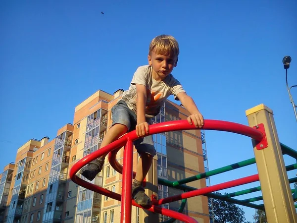 Blonďatý chlapec se vyšplhal po kovové konstrukci na hřišti, rukama se chytil zábradlí a podíval se dolů. Herní zařízení pro dvorek. Cihlový dům a modrá obloha v pozadí. — Stock fotografie