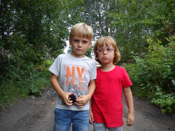 Un niño y una niña son hermano y hermana. Los niños con cabello rubio caminan junto a árboles y arbustos. Chica con gafas y una camiseta roja. Niños posando sobre un fondo de una carretera rural y vegetación —  Fotos de Stock