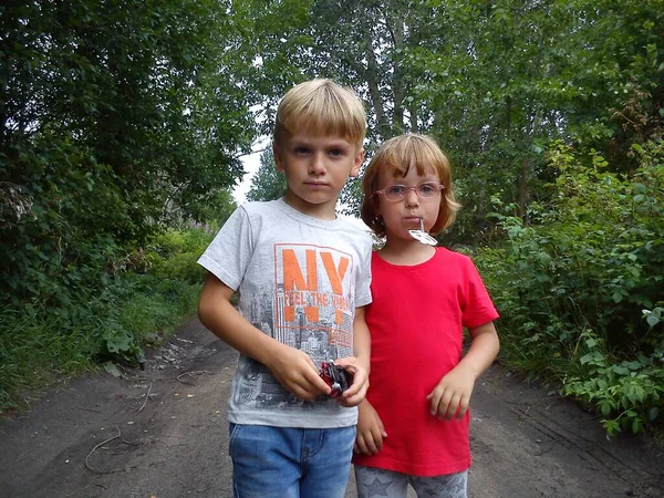 Un niño y una niña son hermano y hermana. Los niños con cabello rubio caminan junto a árboles y arbustos. Chica con gafas y una camiseta roja. Niños posando sobre un fondo de una carretera rural y vegetación —  Fotos de Stock