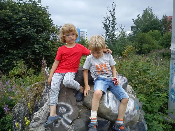 Ένα αγόρι και ένα κορίτσι είναι αδέρφια. Παιδιά με ξανθά μαλλιά περπατούν δίπλα σε δέντρα και θάμνους. Κορίτσι με γυαλιά και κόκκινο μπλουζάκι. Τα παιδιά σκαρφάλωσαν σε μια μεγάλη θέση - λιθόστρωτη — Φωτογραφία Αρχείου
