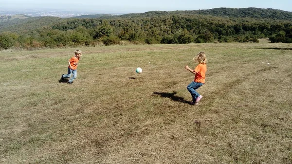 Chlapec a dívka s blond vlasy, oblečeni v oranžových tričkách a klasických džínách, hrát míč na horské louce. Časně teplý podzim. V pozadí horizont a vegetace — Stock fotografie