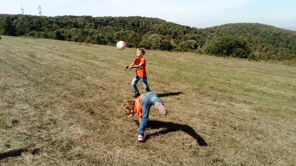 Chlapec a dívka s blond vlasy, oblečeni v oranžových tričkách a klasických džínách, hrát míč na horské louce. Časně teplý podzim. Dívka se překlopí přes hlavu a opře se o ruce — Stock fotografie