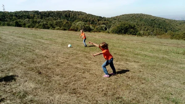 Мальчик и девочка с светлыми волосами, одетые в оранжевые футболки и классические джинсы, играют в мяч на горном лугу. Ранняя теплая осень. Девушка падает на голову, опираясь на руки. . — стоковое фото